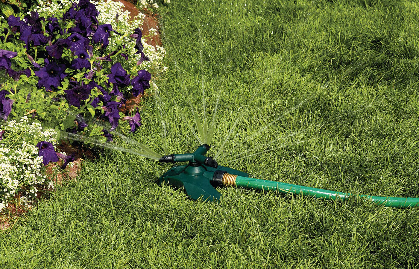 Green and black 3-arm plastic revolving sprinkler on plastic base spraying water on grass and flowers.