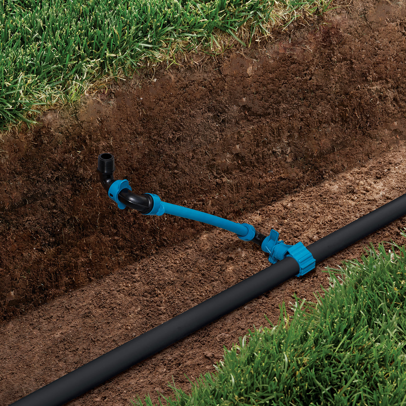 A blue and black drip irrigation system shown installed in a trench outdoors, with grass and soil visible around it.