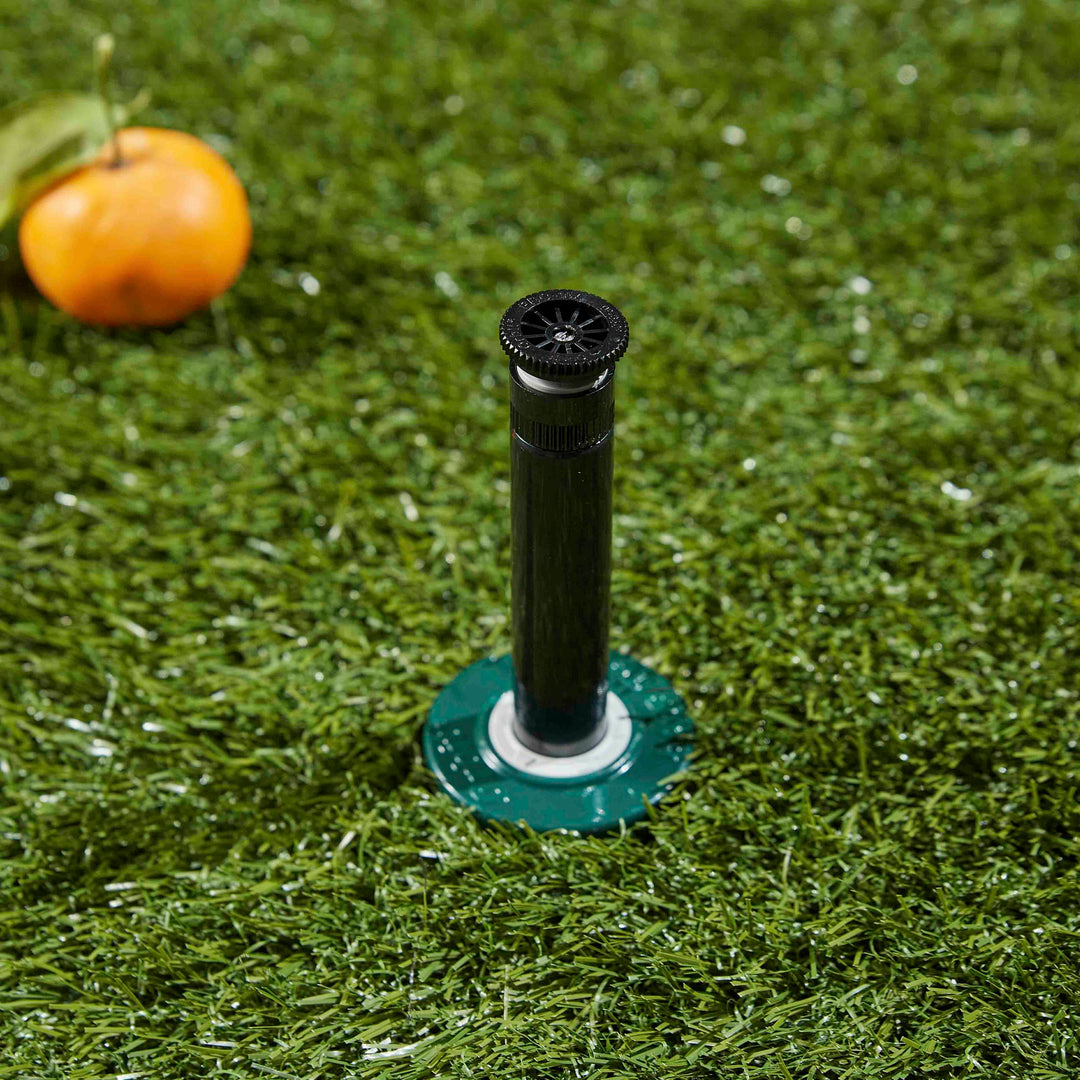 Close up of an extended riser stem on a professional hard top pop-up spray head sprinkler with adjustable nozzle. Installed in ground with grass and a tangerine lying beside it.
